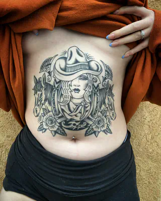 Black and grey tattoo across the stomach of a woman in a cowboy hat framed by flowers and filigree