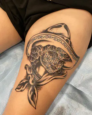 Black and grey tattoo of a figure in a wide sombrero with a sacred geometry face and a knotted bandana, on a thigh