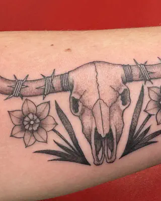 Black and grey steer skull wrapped in barbed wire between two daffodils, on a forearm