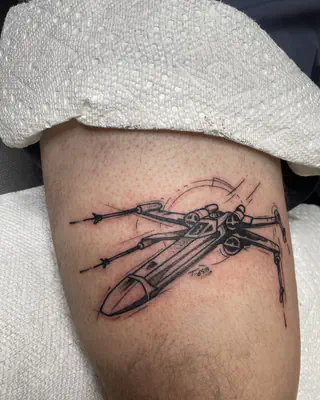 Black and grey line tattoo of an X-wing starfighter, on a calf