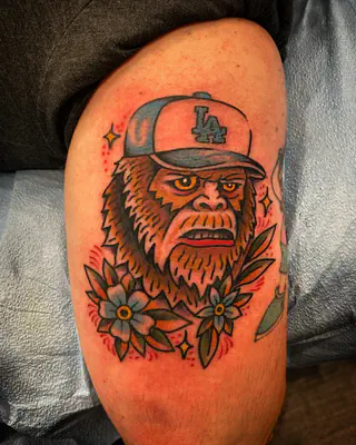 Color tattoo of Bigfoot in a Los Angeles baseball cap above two flowers, on a thigh