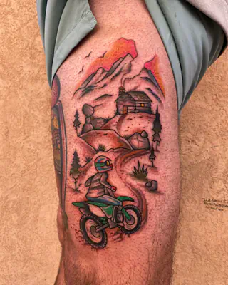 Color tattoo of a dirt bike rider on a trail below a mountain cabin and pines at sunset, on a thigh