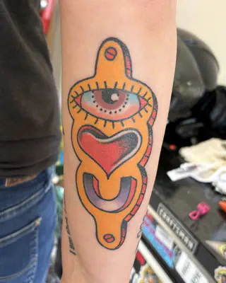 Color tattoo of an eye above a red heart inside an ornate yellow frame, on a forearm
