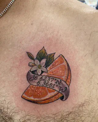 Color tattoo of an orange slice and a white orange blossom wrapped in a banner of map coordinates, on a chest