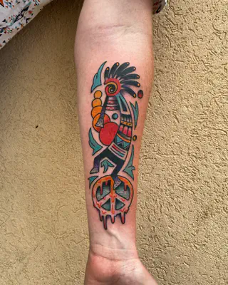 Color tattoo of a dancing Southwest figure in a feathered headdress above a peace sign, on a forearm