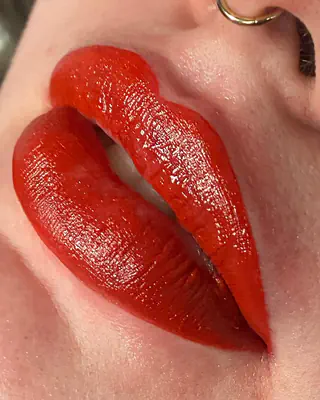 Cosmetic lip tattoo in a bold, glossy red with a crisp lip line, close up beside a gold nose ring