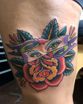 Neo traditional color tattoo of a red rose and green leaves beneath a pair of eyes framed in bold color, on a thigh