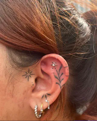 Fine line star and botanical tattoos beside an ear with gold hoop and stud piercings