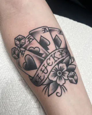 Traditional black and grey hand of aces with dice, a flower, and a Lucky banner, on a forearm